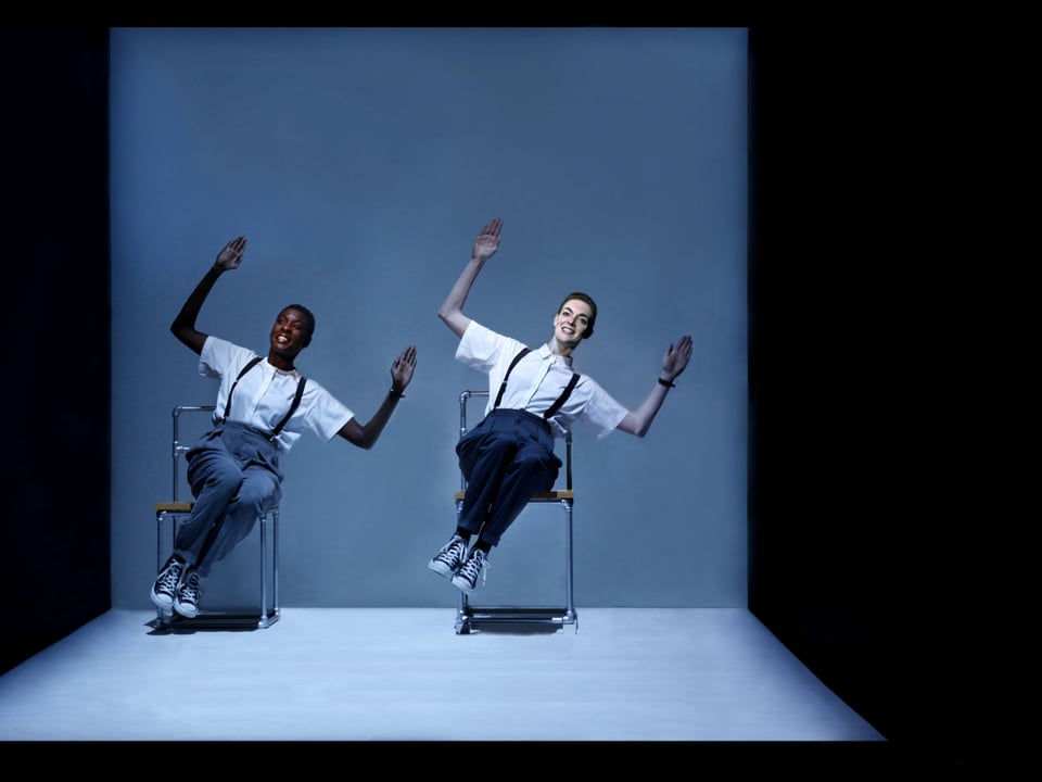 Zwei Tänzer sitzen auf Stühlen und ziegen mit beiden Armen nach rechts oben.